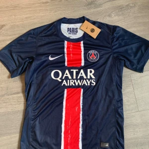 PSG matchtröja  - Paris Saint-Germain fotbollströja från Nike i marinblått med en bred röd och vit rand mitt på bröstet. Tröjan har korta ärmar, rund hals och PSG-logga på bröstet. Materialet är lätt och andas, perfekt för träning eller match. Qatar Airways sponsortryck på framsidan. Storleken är medium. Skriv för minsta fråga