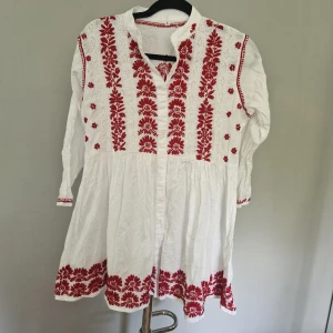 Vit kortklänning med röd brodyr - Superfin vit kortklänning med långa ärmar och röd broderad blommig design framtill, på ärmar och längs nederkanten. Klänningen har v-ringning och är gjord i bomull, perfekt för en somrig och bohemisk look.