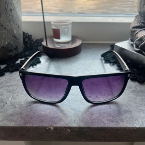Svarta solglasögon med lila tonade färger en copy av RAY BANDS - Snygga solglasögon med svart båge och stora, fyrkantiga linser som är lila tonade. Riktigt clean och modern look, perfekt för dig som vill sticka ut lite extra. Bågarna är i plast och har en klassisk streetstyle-vibe.