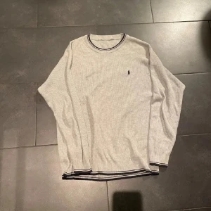 Ljusgrå långärmad tröja från Polo Ralph Lauren - Ljusgrå långärmad tröja från Polo Ralph Lauren med diskret ribbad struktur. Tröjan har mörka kontrastlinjer vid halsringning och nederkant samt den klassiska broderade loggan på bröstet. Perfekt för en clean och stilren look.