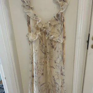 H&M klänning -  långklänning med blommigt mönster i ljusa beige och lila toner. Klänningen har volangdetaljer längs ringningen och axelbanden, samt en luftig och flowy passform. 