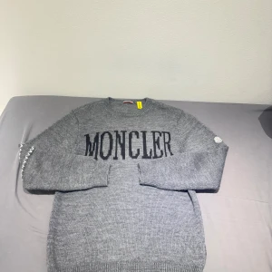 Grå stickad Moncler tröja - Snygg grå stickad tröja från Moncler med svart logotyp över bröstet och coola vita detaljer på ena ärmen. Tröjan har rund halsringning och långa ärmar. Tillverkad i mjuk bomull som känns skön mot huden. Perfekt för dig som gillar stilrena och exklusiva plagg.(nypris 10000) 