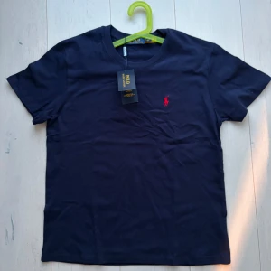 Mörkblå t-shirt från Polo Ralph Lauren - Snygg mörkblå t-shirt från Polo Ralph Lauren med klassisk röd broderad logga på bröstet. T-shirten är i mjuk bomull och har rund hals och korta ärmar. Perfekt för en clean och stilren look.