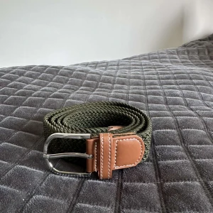 Flätat grönt bälte med skinn-detaljer - Snyggt flätat bälte i mörkgrönt med bruna skinn-detaljer och silvrigt spänne. Bältet har en stilren look med vit kontrastsöm på skinnet och passar perfekt till jeans eller chinos. Flexibel och bekväm modell som ger en cool touch till outfiten.