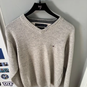 Grå v-ringad ulltröja Tommy Hilfiger - Snygg ljusgrå v-ringad tröja från Tommy Hilfiger i mjuk lammull. Klassisk design med diskret logga på bröstet och ribbade muddar. Perfekt för dig som gillar stilrena och bekväma plagg med premiumkänsla.