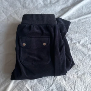 Svarta mjukisbyxor från Juicy Couture - Mysiga svarta mjukisbyxor från Juicy Couture med bred resår och snörning i midjan. Byxorna har stora fickor med knappar bak och två fickor fram. Materialet är mjukt och sammet-liknande, perfekt för chill dagar. En del av de två snörerna har gått av. 