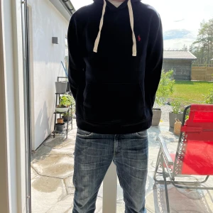 Svart hoodie från Polo Ralph Lauren - Tja säljer marin blå hoodie från Polo Ralph Lauren med klassisk röd logga på bröstet. Tröjan har vita snören i huvan och en stor magficka framtill. Perfekt för dig som gillar en enkel men bra till en stilig look för sommaren! Inga defekter på tröjan om ni har frågor eller funderingar är det bara att höra av er priset är inte hugget i sten! 