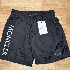 Svarta Moncler shorts med logga - Svarta shorts från Moncler med stor vit logga längs ena benet och broderad patch. De har elastisk midja med snörning och är tillverkade i lätt, glansig polyester med meshfoder. Perfekta för sommaren och chill dagar.