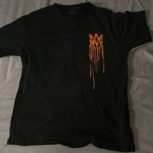 Svart Amiri t-shirt med orange tryck - Behöver bara strykas! Svart t-shirt från Amiri med orange droppande logotyp på bröstet och stort AMIRI-tryck på ryggen. Klassisk passform och rund hals. Materialet känns mjukt och skönt, perfekt för dig som gillar streetwear med edge.