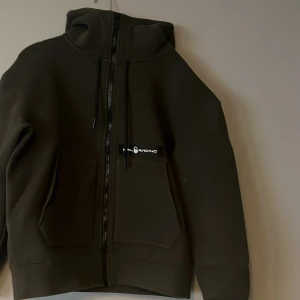 Mörkgrön hoodie från Sail Racing - Säljer en mörkgrön hoodie från Sail Racing med dragkedja framtill, huva med snörning och två stora fickor. Tröjan har en stilren look och logga på bröstet. Perfekt för dig som gillar sportig och clean stil.