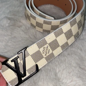 Louis Vuitton rutig bälte med LV-spänne - Snyggt bälte från Louis Vuitton med klassiskt rutigt mönster i beige och grått. Bältet har en stor svart LV-logga som spänne och vita kanter. Tillverkat i skinn med exklusiv känsla, perfekt för att lyfta vilken outfit som helst.