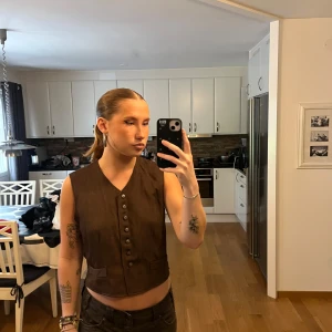 Brun väst med knappar - Säljer en vintage brun väst med snygg v-ringning och raka axlar. Västen har flera knappar framtill och två dekorativa fickor. Perfekt att styla över en t-shirt eller skjorta för en cool look.