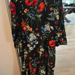 Svart blommig långklänning med knappar - Svart långklänning med färgglatt blommönster i rött, blått, vitt och orange. Klänningen har långa ärmar och knappar framtill. Perfekt för dig som gillar en färgstark och trendig look.