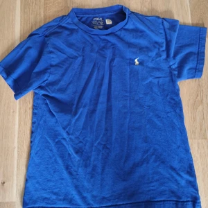 Blå t-shirt från Polo Ralph Lauren - Klassisk blå t-shirt från Polo Ralph Lauren med broderad logga på bröstet. T-shirten har rund halsringning och korta ärmar. Tillverkad i mjuk bomull som är skön att bära hela dagen. Perfekt för en avslappnad och stilren look.