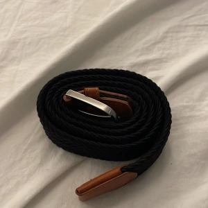 Svart flätat bälte med läderdetaljer - Stilrent svart flätat bälte med bruna läderdetaljer och silverfärgat spänne. Bältet har en twistad design som ger en cool och modern känsla. Perfekt för att lyfta din outfit med en snygg accessoar.