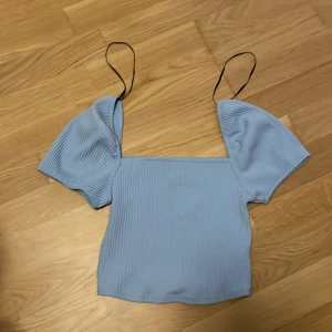 Ljusblå ribbad topp från H&M - Supersöt ljusblå topp från H&M med ribbad struktur och puffiga korta ärmar. Toppen har smala svarta axelband och fyrkantig halsringning. Perfekt för dig som gillar en trendig och feminin stil.