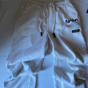 Vita NASA mjukisbyxor med tryck - Vita mjukisbyxor med NASA-tryck på ena benet. Byxorna har resår i midjan och snörning för extra komfort. Tillverkade i mjukt bomullsmaterial, perfekta för en chill och avslappnad stil.