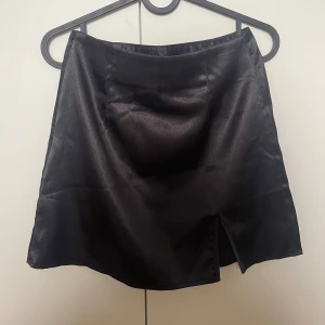 Svart kortkjol med slits - Snygg svart kortkjol med en liten slits framtill. Kjolen har en stilren och enkel design med lätt glansigt tyg. 