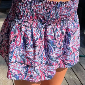 Färgglad kortkjol med paisleymönster - Superfin kortkjol med färgstarkt paisleymönster i blått, rött och vitt. Kjolen har bred smock i midjan för skön passform och volangig nederdel som ger en söt och luftig känsla. Perfekt för varma dagar när du vill sticka ut lite extra. Den passar xxs-xs skulle jag säga