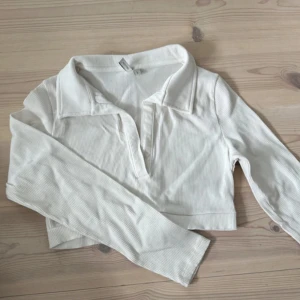 Vit ribbad croppad pikétröja från Nelly - Snygg croppad pikétröja från Nelly i vit ribbad bomull. Tröjan har lång ärm, klassisk krage och djupare v-ringning framtill. Perfekt till jeans eller kjol för en clean och fräsch look.