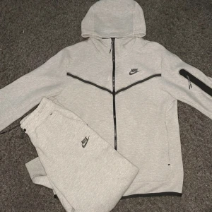 Nike tech fleece kofta  - Nike tracksuit i ljusgrått med svart dragkedja och detaljer. Hoodien har huva, svarta linjer över bröstet och en praktisk ficka med dragkedja på ärmen. Byxorna har bara tröjan finns kvar  resår i midjan och Nike-logga på låret. Perfekt för chill eller träning. Står Xs men passar mer som en M 