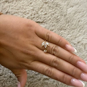 Guldig ring med blommig kristalldesign - Superfin guldig ring med öppen design och gnistrande kristaller formade som en blomma. Ringen har flera klara stenar i olika former som ger en lyxig och unik look. Perfekt för dig som vill sticka ut med något extra snyggt på fingret.