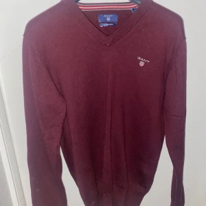 Vinröd v-ringad tröja från Gant - Vinröd v-ringad tröja från Gant med broderad logga på bröstet. Tröjan är långärmad och har en klassisk passform. Tillverkad i en mjuk blandning av bomull och ull, perfekt för lager-på-lager under kyliga dagar.