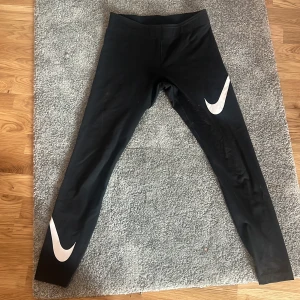 Svarta Nike leggings med logga - Svarta leggings från Nike med stora vita swoosh-loggor på båda benen. Tight passform och stretchigt material som passar perfekt till träning eller chill. Klassisk och sportig look.