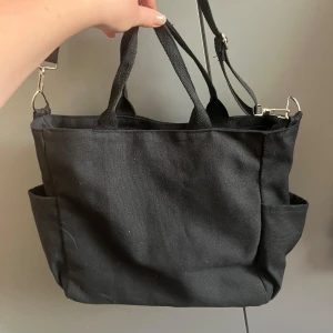 Svart axelväska i bomull - En stilren svart axelväska i bomull med både handtag och justerbar axelrem. Väskan har flera praktiska ytterfickor och stängs med dragkedja upptill. Perfekt för dig som vill ha en enkel och rymlig väska till vardagen.