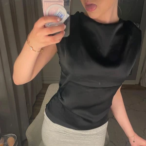 Svart satinblus - Snygg svart blus i satin med markerade axlar och ärmlös design. Blusen har en elegant, slät yta och rund halsringning. Perfekt för att lyfta vilken outfit som helst med en stilren touch.