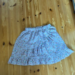 Blommig volangkjol - Supersöt kort kjol i ljuslila med små blommor. Kjolen har volanger och resår i midjan. I storlek 140 men är strechig så passar någon som vanligtvis bär XS. Kort och fin sommarkjol. Fint skick! KAN ABSOLUT GÅ NER I PRIS VID SNABB AFFÄR ELLER BUNDLE!
