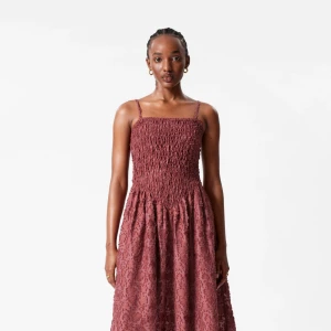 Gammelrosa långklänning med smockliv - Superfin gammelrosa långklänning med smala axelband och smockat liv. Kjolen är lätt utställd och har ett spetsblommigt mönster i tyget. Perfekt för dig som vill ha en romantisk och trendig look.