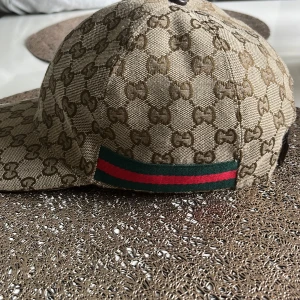 Gucci keps med GG-mönster och röd/grön detalj - Snygg keps från Gucci i beige med klassiskt GG-monogram över hela kepsen. Kepsen har en ikonisk röd och grön rand på sidan och justerbar rem baktill i brunt. Tillverkad i bomull och polyester, perfekt för dig som vill sticka ut med lyxig streetstyle.