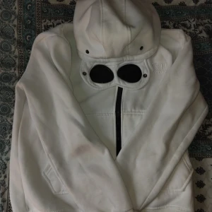 Vit gogglejacka från C.P. Company - Säljer en vit gogglejacka från C.P. Company med ikoniska svarta glasögon i huvan och broderad logga på bröstet. Jackan har dragkedja framtill och är tillverkad i ett mjukt bomullsmaterial. Perfekt för dig som vill sticka ut med en unik streetwear-look.