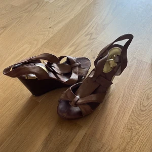 Bruna kilklackade sandaler H&M - Snygga bruna sandaler från H&M med kilklack och öppen tå. Remmar i brunt skinnimitation och guldfärgat spänne. Perfekta för sommarens alla outfits och ger en skön höjd utan att tumma på komforten.