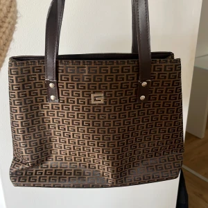 Brun axelväska med G-mönster - Snygg brun axelväska med klassiskt G-mönster över hela väskan. Väskan har två långa handtag i mörkbrunt skinn och flera fack med dragkedja.