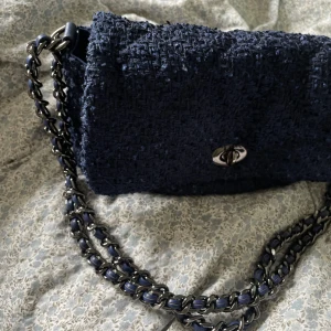  väska becksöndergaard - snygg mörkblå axelväska i tweed med cool kedjerem i metall och blått. Väskan har ett klassiskt lock med vridlås i silverfärg och ett beige innerfoder med dragkedjefack. Perfekt för dig som vill ha en trendig väska till sommaren. 