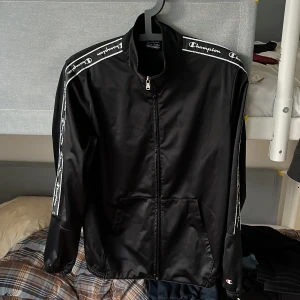 Svart vindjacka från Champion 2XL - Svart vindjacka från Champion i storlek 2XL. Jackan har dragkedja framtill, vita logoband längs ärmarna och två fickor. Materialet är glansigt och lätt, perfekt för en sportig look. Elastiska muddar vid ärmslut och nederkant för extra komfort. 