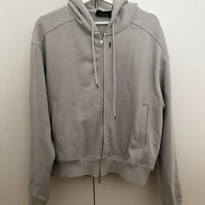 Grå hoodie Eclipse Shelter T.C.G.A - Snygg grå hoodie från Eclipse Shelter med dragkedja och huva. Tryck med texten 'Eclipse - Shelter' och 'T.C.G.A' på ryggen. Mjuk bomullskänsla, långa ärmar och praktiska fickor framtill. Perfekt för en chill och stilren look.