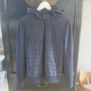 Mörkblå cardigan jacka från J Lindeberg - Snygg mörkblå jacka från JLindeberg med quiltad framsida och rygg, stickade ärmar och muddar. Jackan har luva, hel dragkedja och två fickor med dragkedja. Perfekt för kyliga dagar när du vill ha både stil och komfort. Storlek S