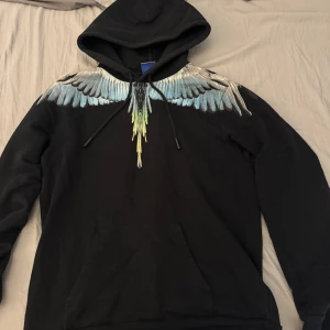 Svart  Marcelo burlon Hoddie med vingar från Marcelo Burlon - Svart Marcelo burlon  hoodie från Marcelo Burlon med ikoniskt vingtryck i blått, vitt och grönt över axlarna. Tröjan har huva med snörning och en stor magficka. 