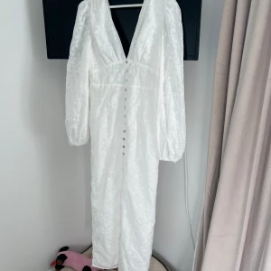 Vit långklänning med broderade blommor - Superfin vit långklänning med djup v-ringning, långa puffärmar och knappar framtill. Klänningen har broderade blommor och blad över hela tyget och är gjord i ett lätt, luftigt material. Perfekt för sommarens alla tillfällen!