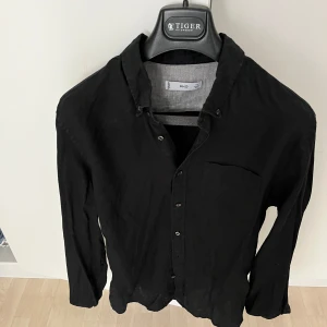 Svart linneskjorta från MNG(mango) - Svart skjorta från mango i klassisk krage och knäppning framtill. Tillverkad i svalt linne. Skjortan har en bröstficka och långa ärmar.