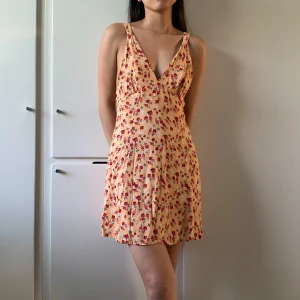 Blommig kort sommarklänning - Söt kort klänning i ljusgul ton med små röda blommor. Djup v-ringning och smala axelband ger en luftig känsla. Axelbandens längd kan justeras bak på ryggen. Dragkedja i sidan. Ser ingen storlek men uppskattas vara XS/S.