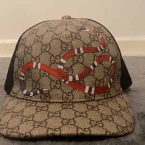 Gucci keps med orm och GG-mönster - Snygg keps från Gucci med klassiskt GG-monogram i beige och brunt, prydd med en röd, vit och svart orm framtill. Baksidan är i svart mesh för extra ventilation. Märkeslapp och detaljer i hög kvalitet. Perfekt för dig som vill sticka ut med lyxig streetstyle.