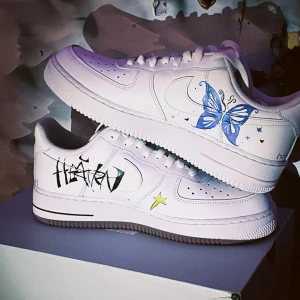 Nike Air Force 1  - Unika vita Nike Air Force 1 sneakers med handmålade motiv som fjäril, graffiti-text, stjärnor och tecknade figurer. Skorna har klassisk låg siluett, snörning och platt sula. Perfekta för dig som vill sticka ut med en kreativ och personlig stil.