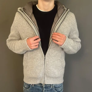 Gran sasso stickad jacka med huva och dunfoder - Snygg grå stickad jacka med huva och quiltat dunfoder på insidan. Jackan har dragkedja framtill, ribbade muddar och två fickor. Perfekt för kyliga dagar när du vill vara både varm och stilren.
