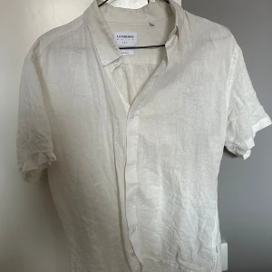 Vit linneskjorta från Lindbergh - Snygg vit skjorta från Lindbergh i svalt linne. Skjortan har korta ärmar, klassisk krage och knäppning framtill. Perfekt för varma dagar när du vill ha en clean och fräsch look.