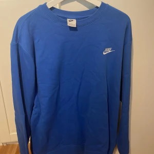 Blå crewneck från Nike - Snygg blå crewneck från Nike med klassisk logga broderad i vitt på bröstet. i har rund halsringning och långa ärmar, perfekt för en chill och sportig stil. Tillverkad i bomullsmix som känns skön mot huden.