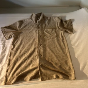 Beige kortärmad frottéskjorta från Kappahl - Kortärmad beige skjorta från Kappahl med knappar framtill och en bröstficka. Skjortan har en mjuk frottéliknande känsla och avslappnad passform, perfekt för en chill look. Enkel och stilren design som funkar till många outfits. Helt oanvänd.